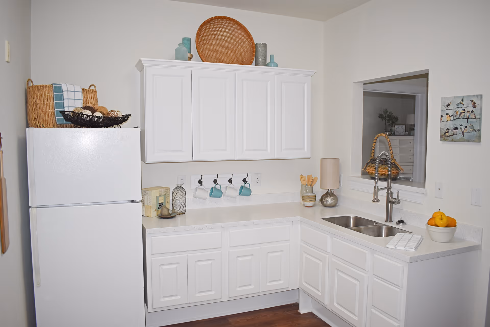 A clean and bright kitchen with white cabinets and countertops. There is a white refrigerator on the left with a basket and decorative items on top. The countertop has a few decorative items including a lamp, a bowl of fruit, and some kitchen utensils. A double sink with a modern faucet is on the right side under a pass-through window to another room. The walls are white, and there is a small painting of birds on the right wall.