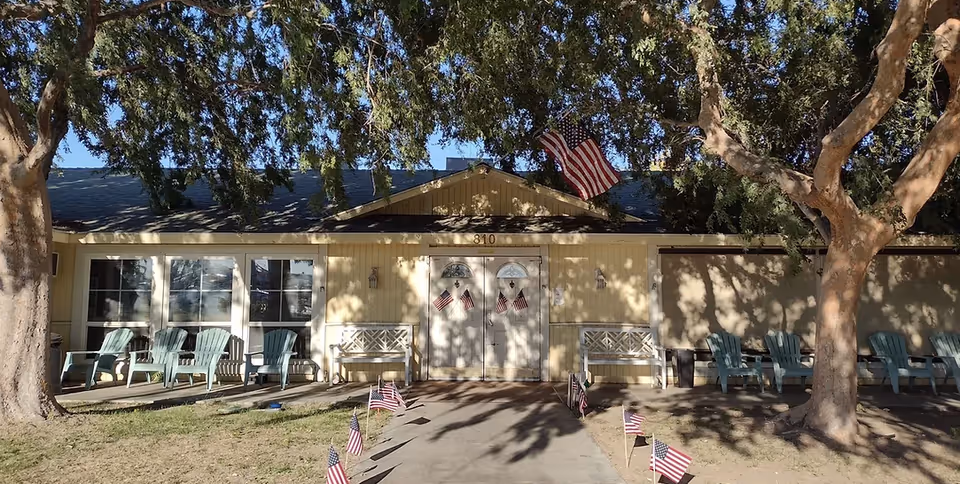 Front exterior view of a single-story assisted living facility building with a yellow facade, two white doors decorated with small American flags, several green chairs and white benches on the porch, large trees providing shade, and a pathway lined with small American flags leading to the entrance.