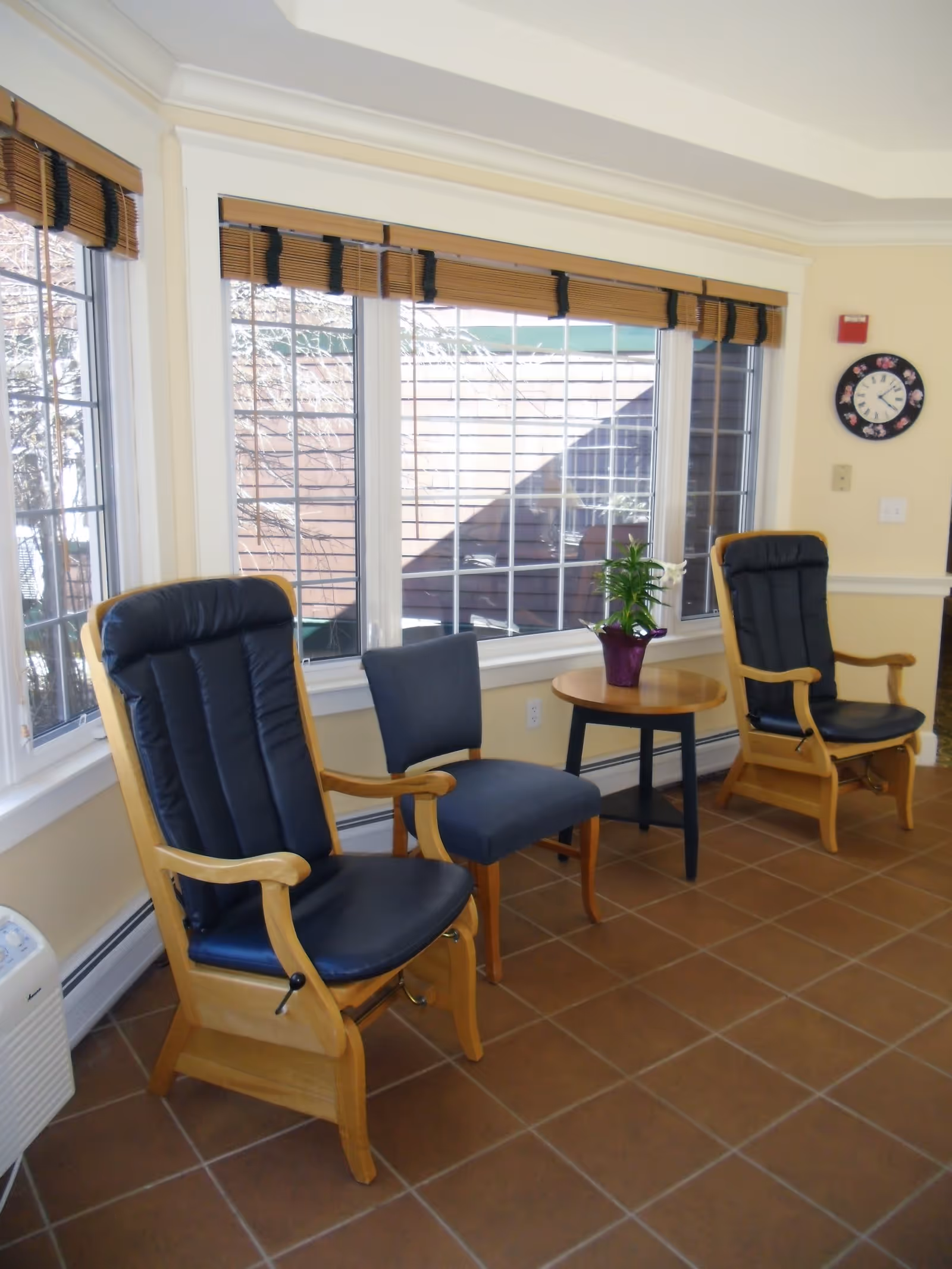 A sunlit seating area with two wooden armchairs featuring black cushions and a smaller blue upholstered chair between them. A round wooden table with a potted plant sits next to the chairs. Large windows with bamboo blinds allow natural light to fill the room, and a wall clock is visible on the beige wall.