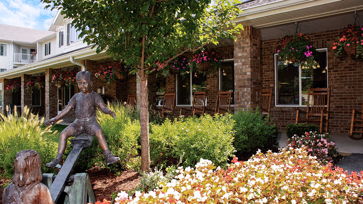 Brick-front porch and landscaped garden with rocking chairs, hanging flower baskets, and a bronze statue of children on a seesaw.
