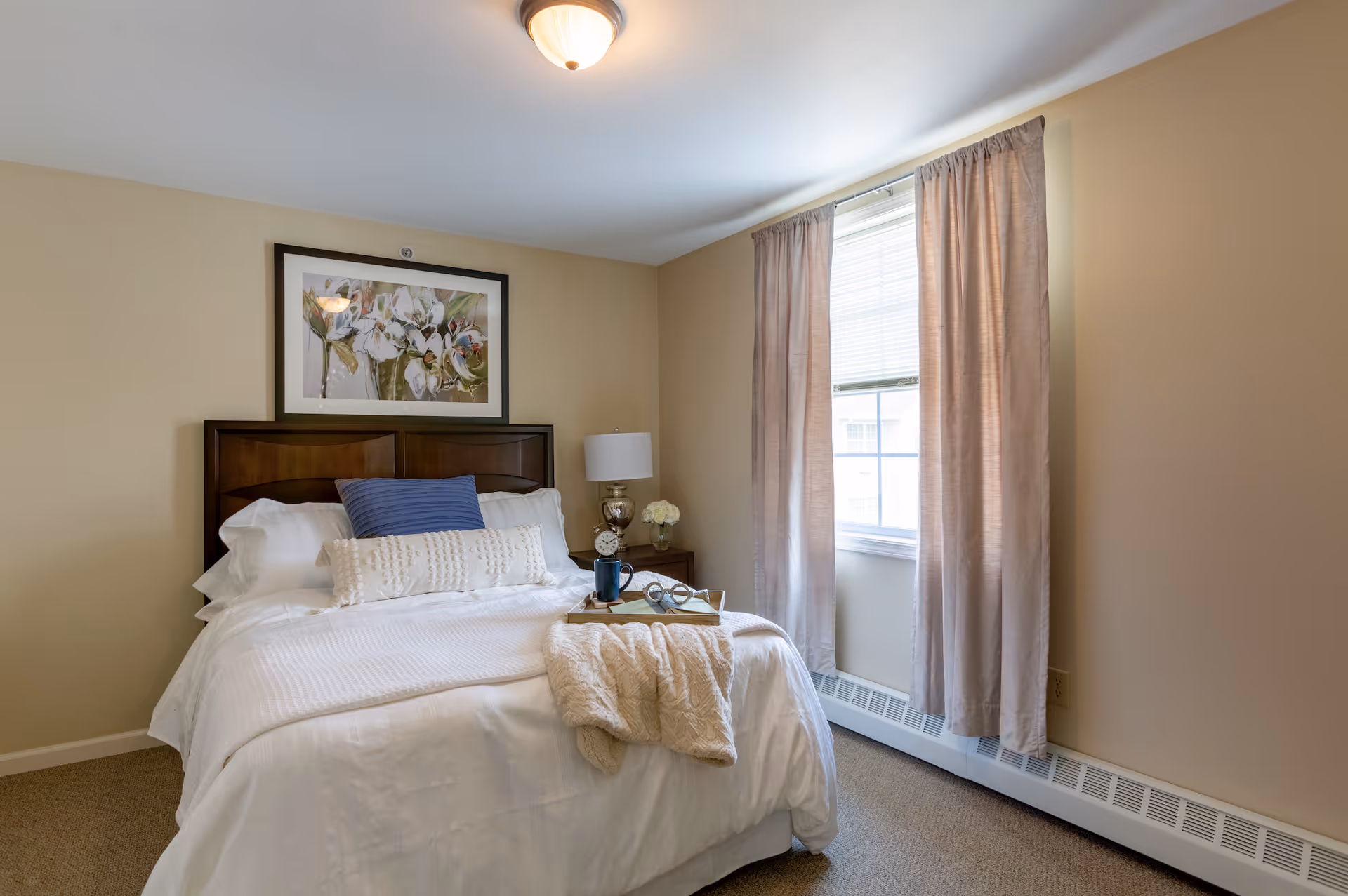 A cozy bedroom with a neatly made bed featuring white bedding, a blue pillow, and a decorative white pillow. A beige throw blanket and a tray with a mug and glasses rest on the bed. There is a wooden headboard, a nightstand with a lamp, an alarm clock, and a small flower vase. A window with beige curtains allows natural light into the room, and a framed floral artwork hangs above the bed.