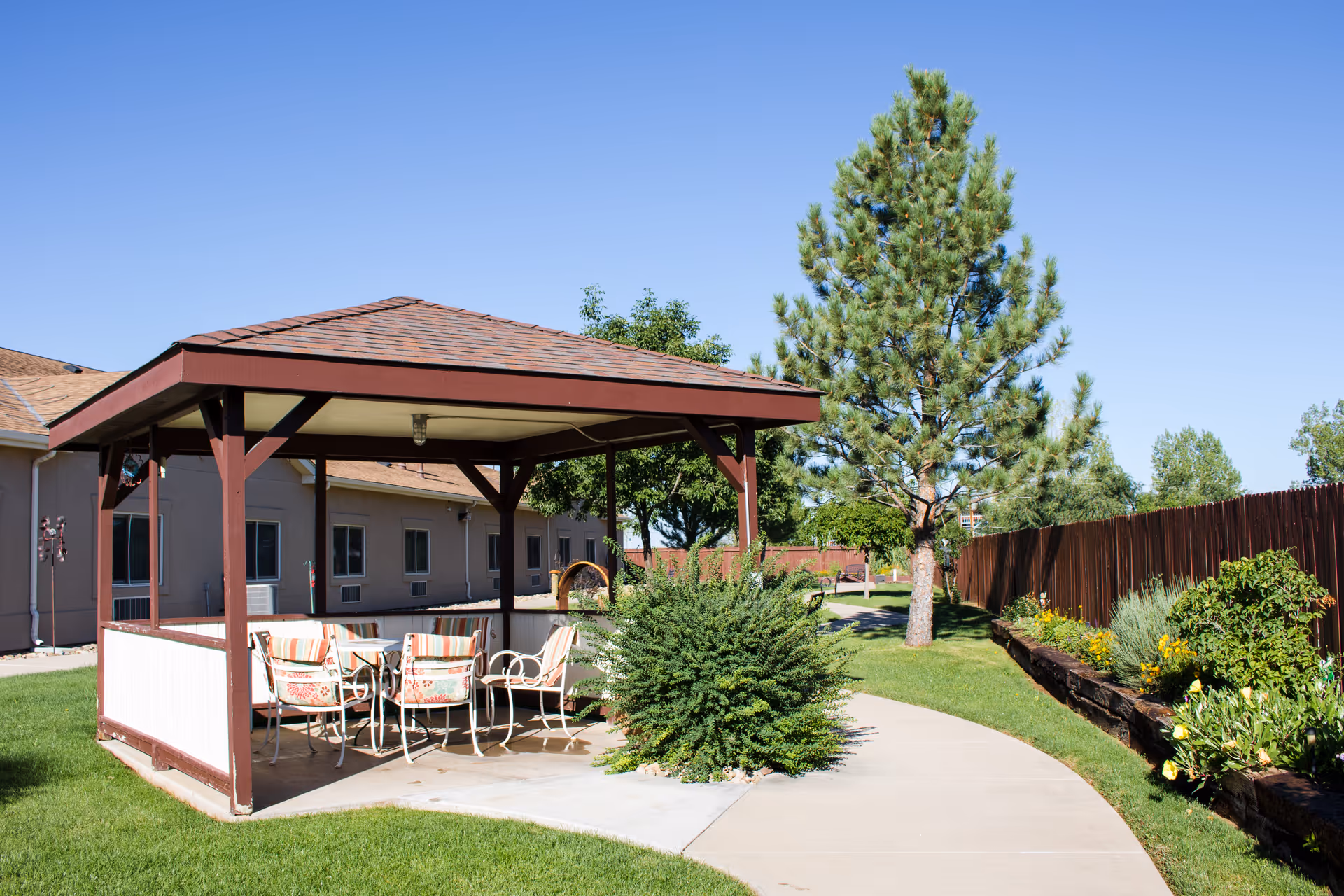 Outdoor gazebo with a table and four chairs with cushions, surrounded by green grass, bushes, trees, and a wooden fence under a clear blue sky at Vista Mesa Assisted Living.