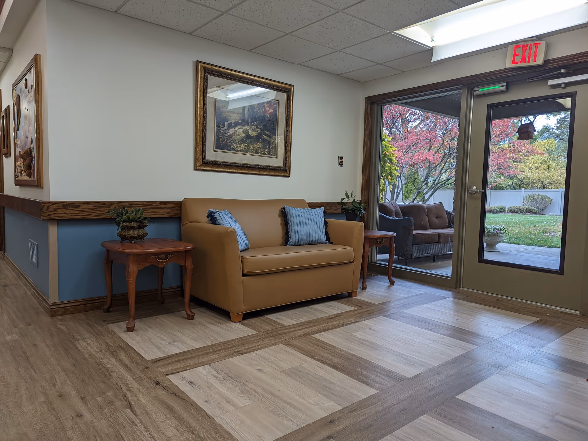 A small lounge with a tan loveseat, side tables and pillows by glass doors opening to a patio and garden.