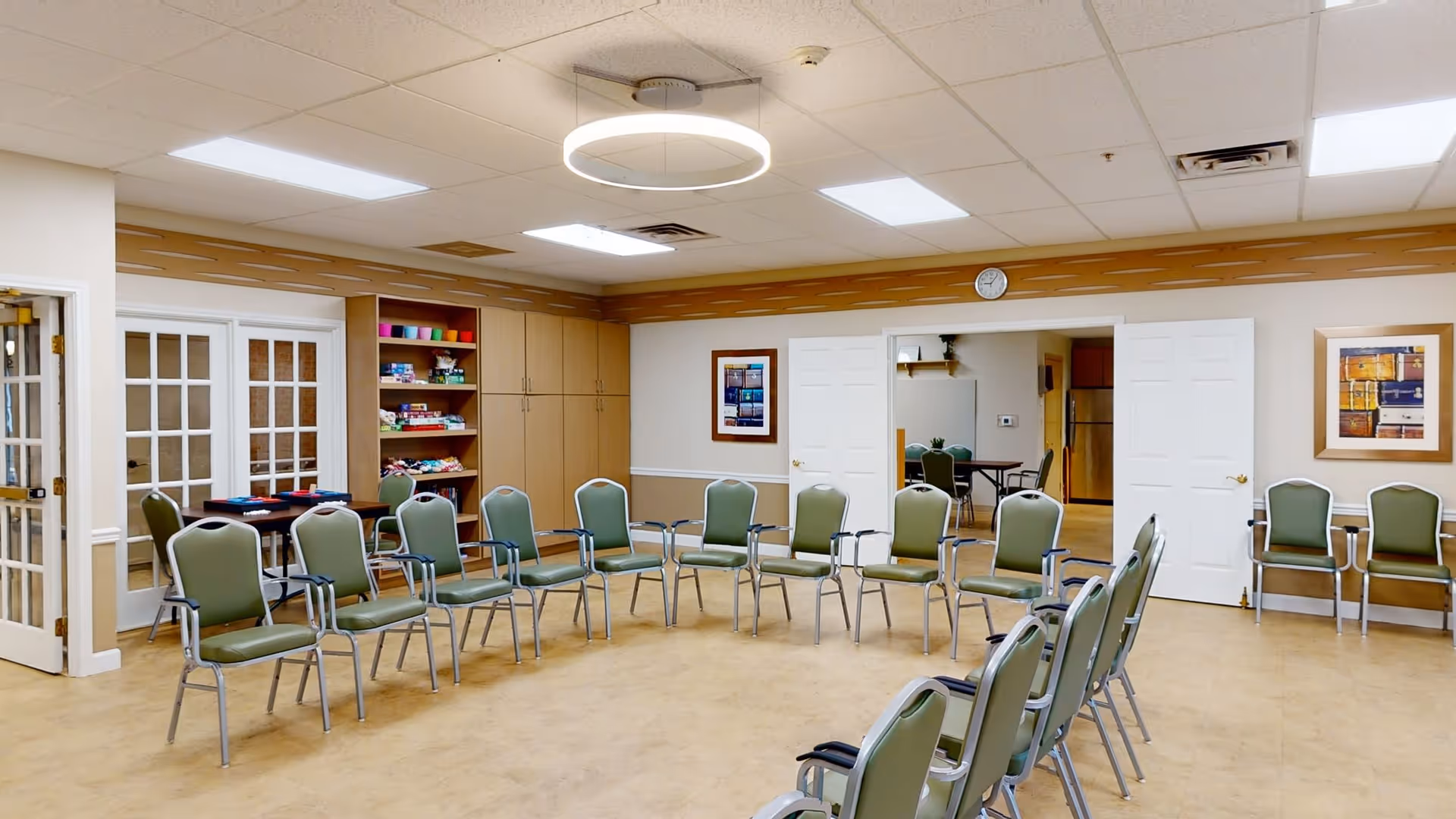 A spacious room with chairs arranged in a large circle, beige walls with wooden trim, a clock above double doors, shelves with colorful items, and framed artwork on the walls. The room has a light-colored floor and a ceiling with fluorescent lights and a circular ceiling light fixture.