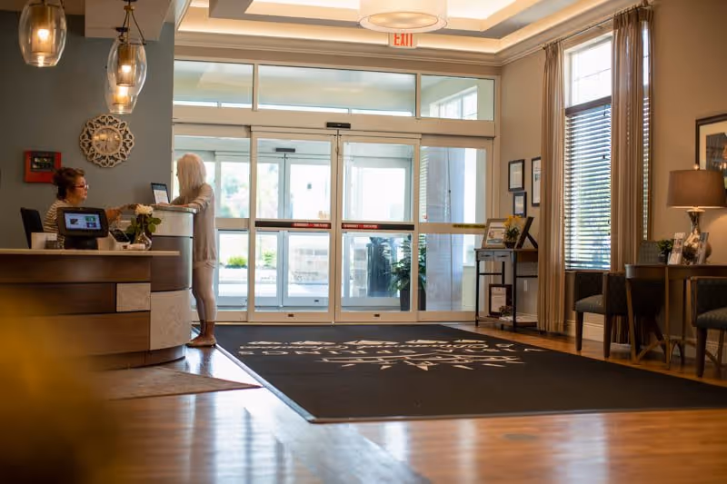 Lobby reception area with a desk where a staff member assists a visitor, automatic glass entrance doors, seating and a large branded floor mat.