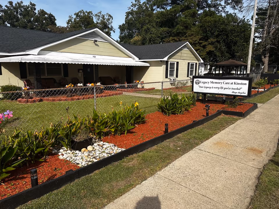 Single-story memory care building with a fenced front yard, landscaped flower beds and a sidewalk sign that reads Legacy Memory Care at Kinston.