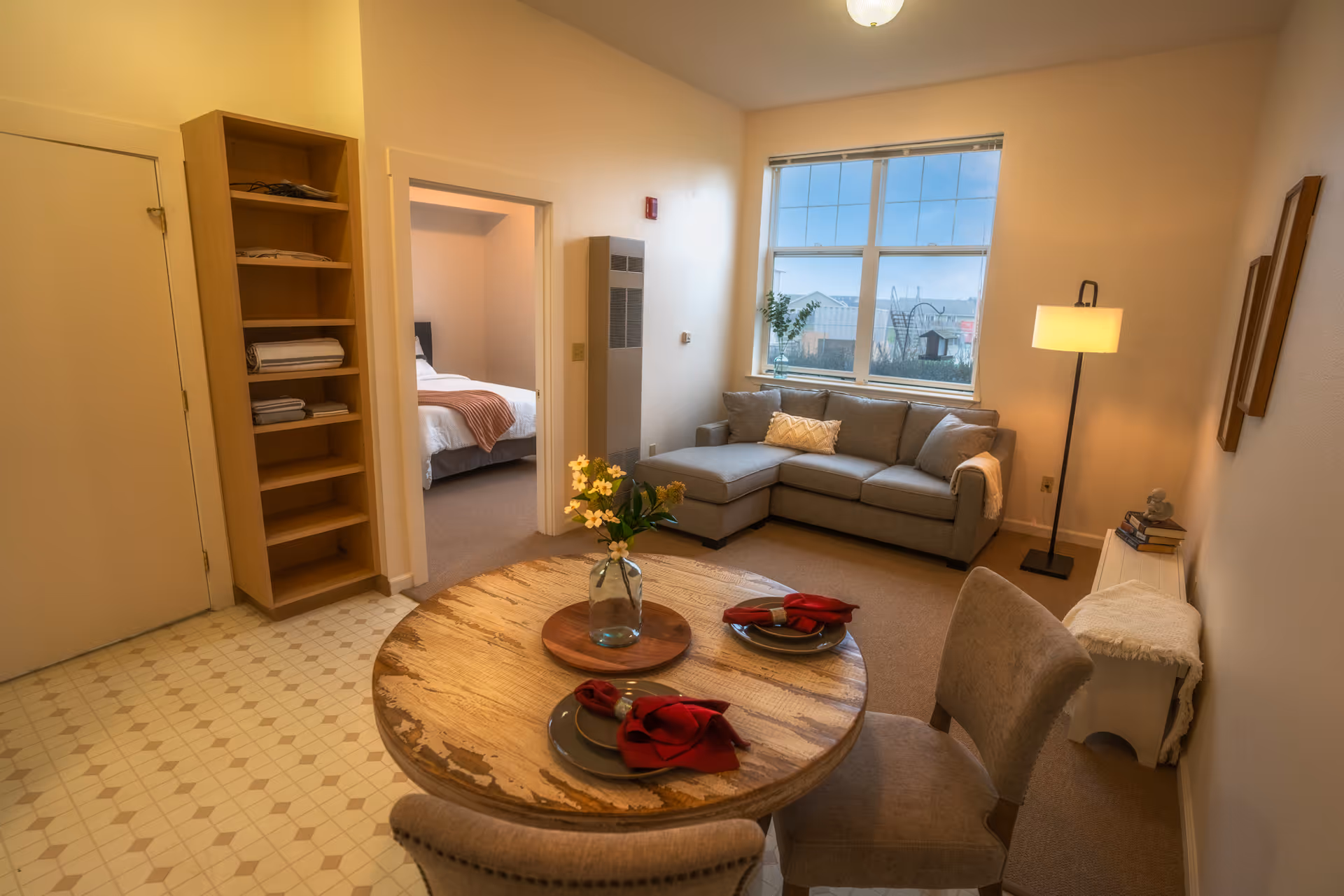 Cozy apartment living area with a round dining table in the foreground, a sectional sofa by a window, and a doorway leading to a bedroom.