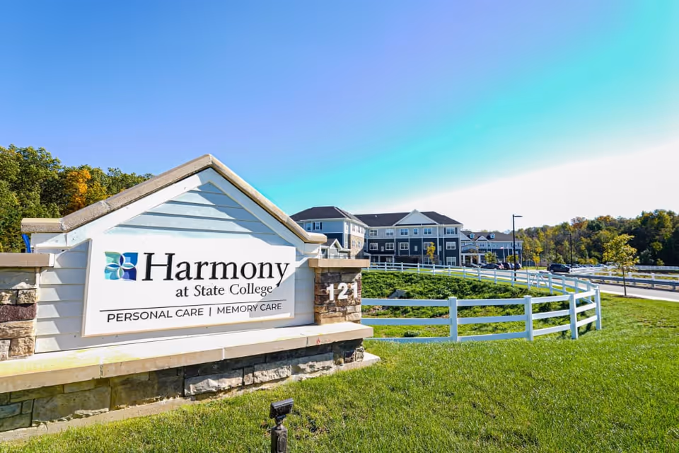 Outdoor view of Harmony at State College senior living facility sign with the building and parking area in the background under a clear blue sky.