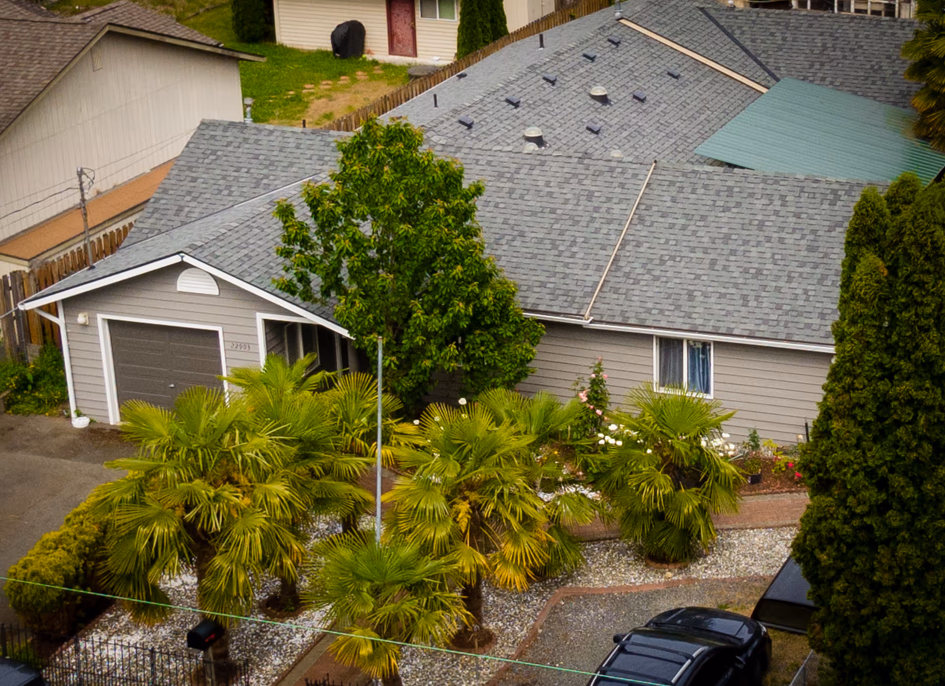 Aerial view of a single-story residential building with a gray shingled roof, a garage, and a small garden area featuring several palm trees and other greenery. A black car is parked in the driveway next to the house.