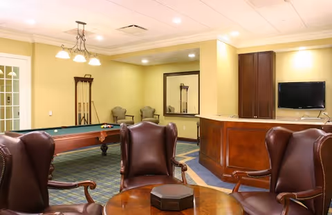 A cozy interior room with a pool table, four leather armchairs arranged around a round wooden table, two additional chairs against the wall, a wall-mounted TV, and warm yellow walls with recessed lighting.