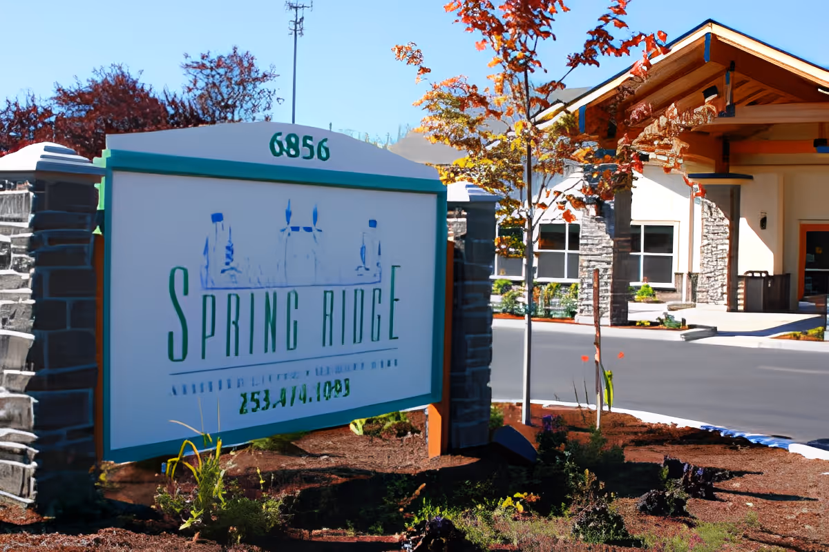 Outdoor view of the entrance to Spring Ridge Assisted Living and Memory Care facility, featuring a large sign with the facility's name and address number 6856, surrounded by landscaping with small plants and trees, and a building with stone and wood architectural details in the background.