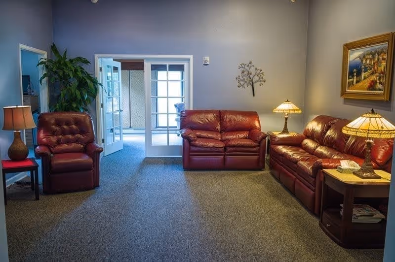 Seating area with red leather sofas and armchairs, side tables and lamps, and glass doors leading to another room.