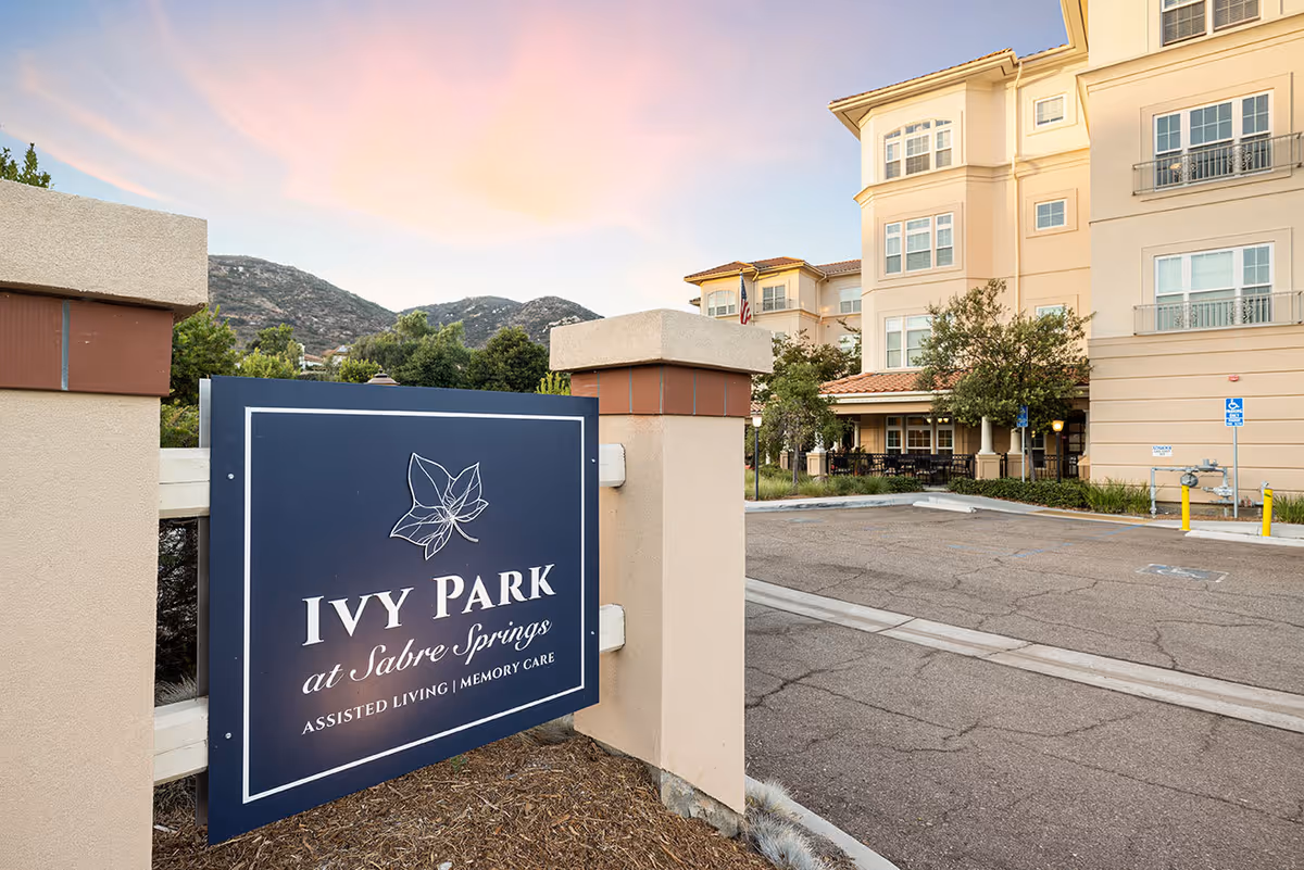 Entrance sign for Ivy Park at Sabre Springs assisted living and memory care facility with the building and parking area visible in the background under a colorful sky at sunset.