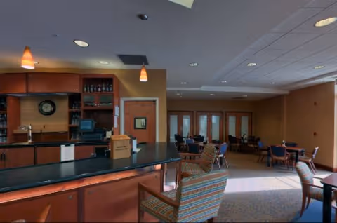 Interior view of a senior living community common area with a bar counter in the foreground, cushioned chairs, and tables arranged for dining or socializing. The room has warm lighting and a carpeted floor with large windows allowing natural light.
