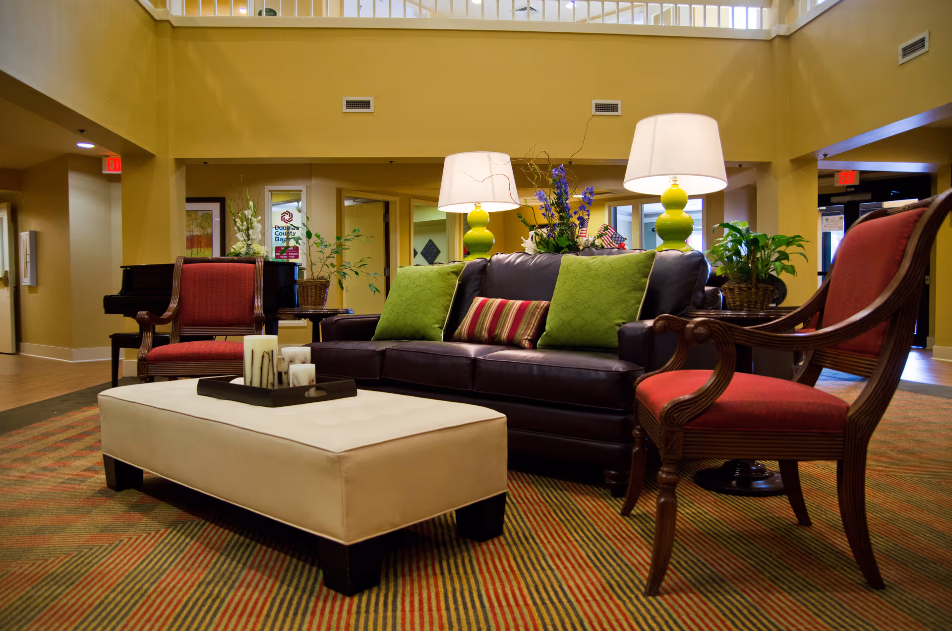 A cozy living room area in a senior living facility featuring a dark leather sofa with green and striped pillows, two wooden armchairs with red cushions, a large beige ottoman with candles on a tray, two green table lamps with white shades, and various plants and flowers. The room has warm yellow walls and a striped carpet, with a piano visible in the background.