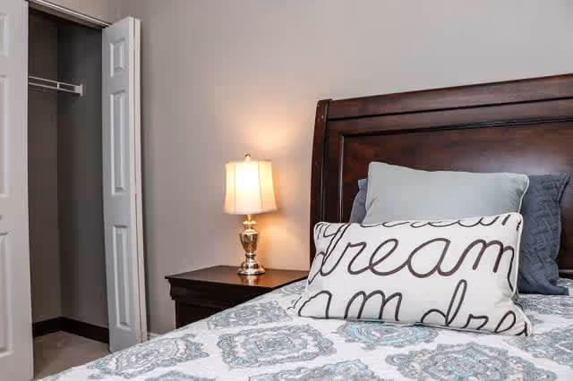 A bedroom with a wooden headboard bed featuring decorative pillows, including one with the word 'dream' written on it. Next to the bed is a wooden nightstand with a lit table lamp. An open closet door is visible in the background.