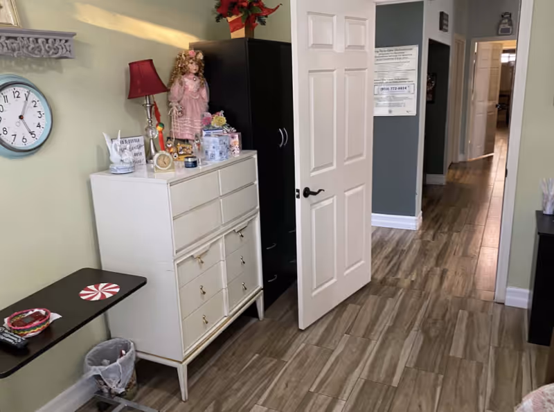 Interior view of a hallway in a senior living facility with a white dresser topped with decorative items including a doll, a red lamp, and various small objects. A white door is open leading to another room, and the floor is covered with wood-patterned tiles. A clock is mounted on a light green wall, and a small table with a remote control and a trash bin underneath is visible.