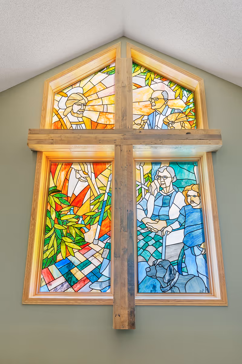 A large stained glass window divided into four sections by a wooden cross. The stained glass depicts various figures including a man with a halo, an elderly man, an elderly woman sitting in a wheelchair, a child, and a dog, surrounded by colorful foliage and geometric patterns.