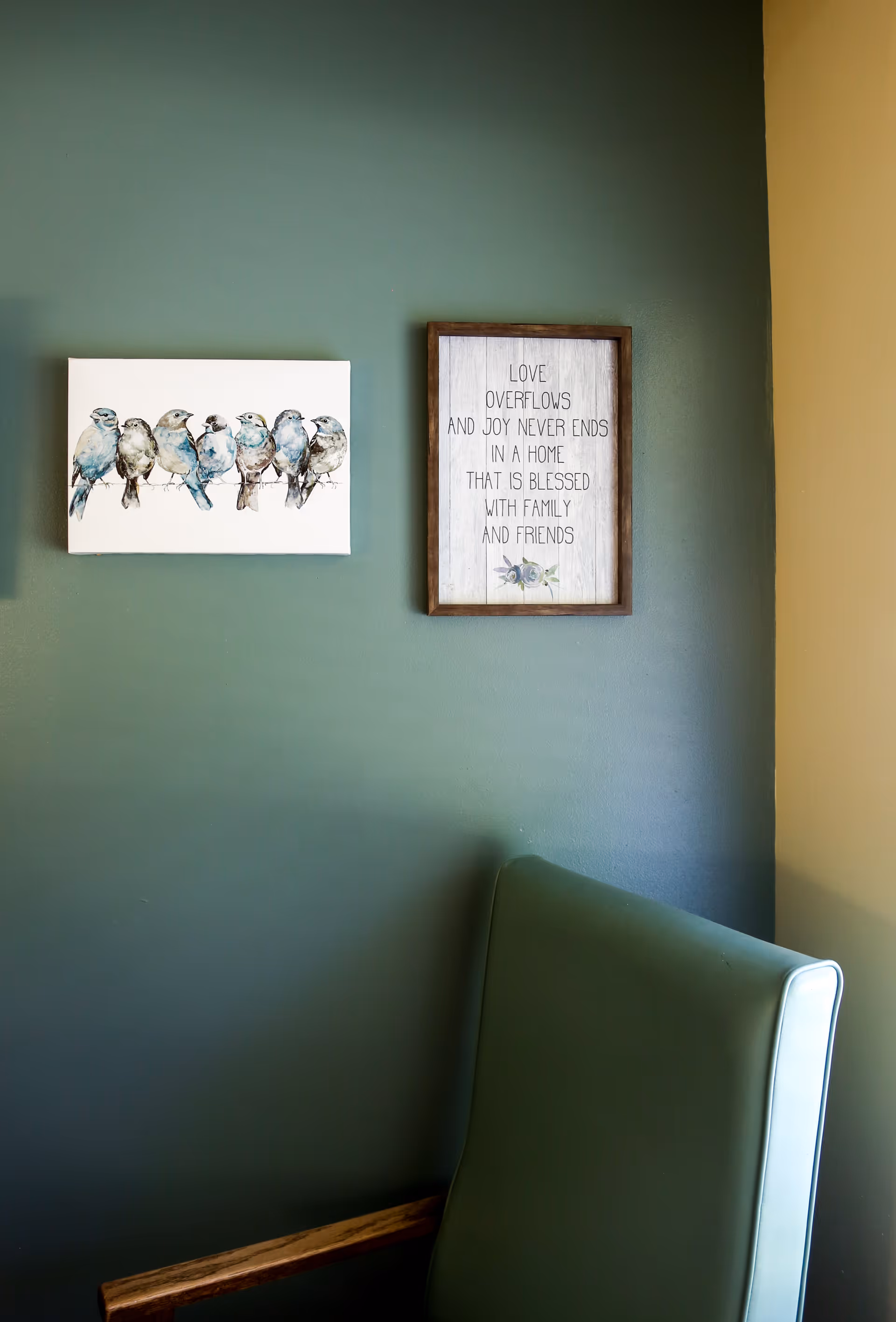 A corner of a room with a green cushioned chair and two framed wall decorations. One frame shows a painting of seven birds sitting on a wire, and the other frame has a quote that reads: 'LOVE OVERFLOWS AND JOY NEVER ENDS IN A HOME THAT IS BLESSED WITH FAMILY AND FRIENDS.' The walls are painted in two colors: green and beige.