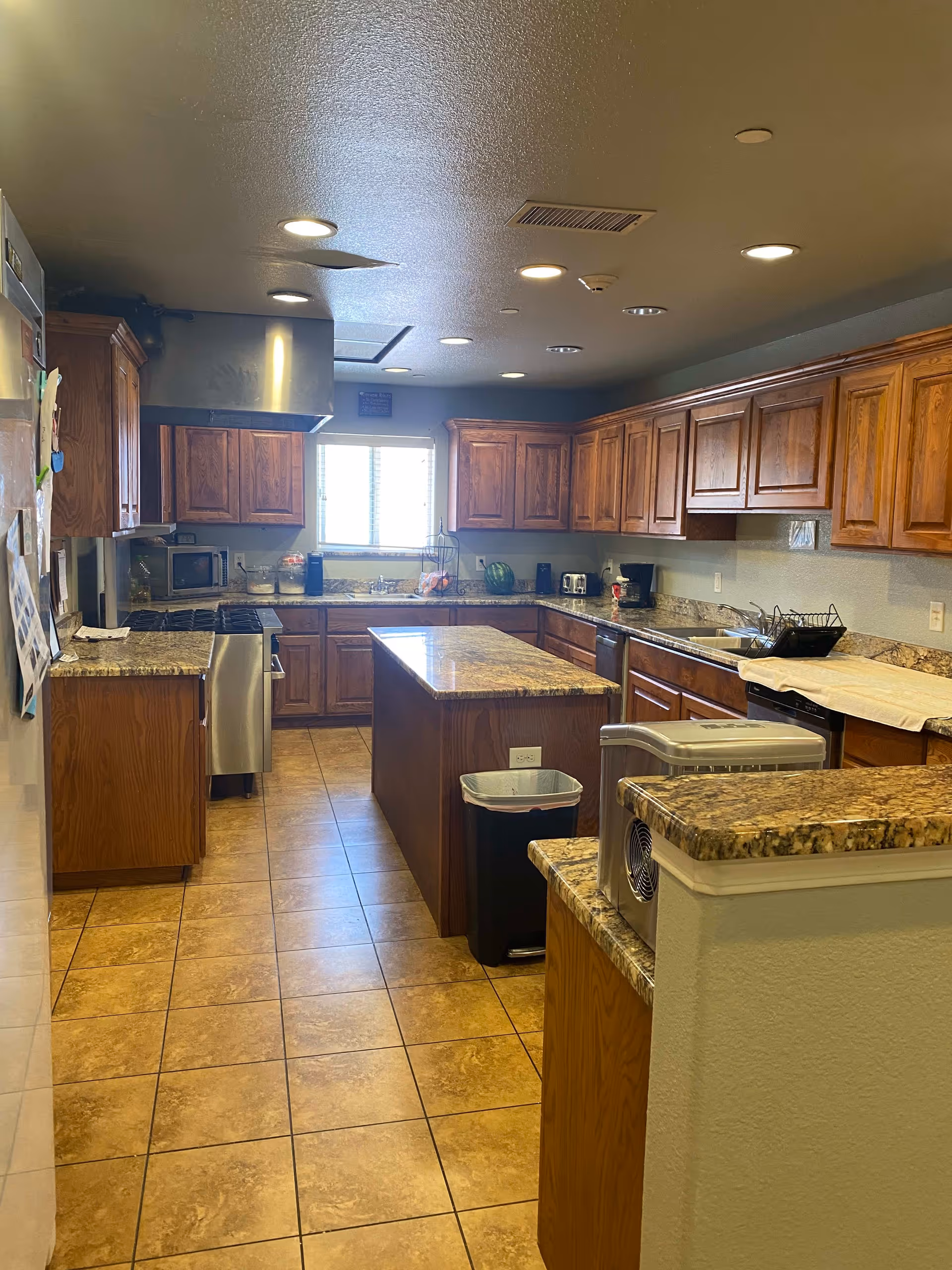A spacious kitchen with wooden cabinets and granite countertops. The kitchen features a central island, stainless steel stove with a range hood, microwave, toaster, coffee maker, and a dishwasher. The floor is tiled, and there is a window above the sink letting in natural light.