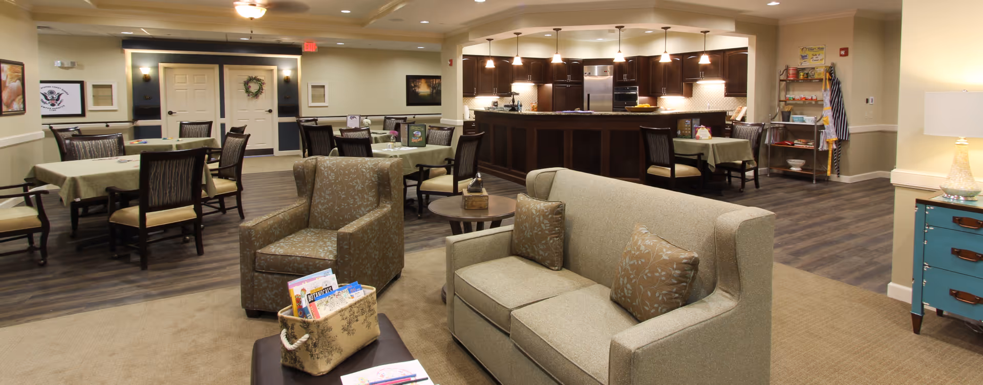 A spacious common area in a senior living facility featuring a seating area with a sofa and armchair, several dining tables with chairs, and a kitchen area with dark wood cabinetry and stainless steel appliances. The room has warm lighting and a cozy, inviting atmosphere.