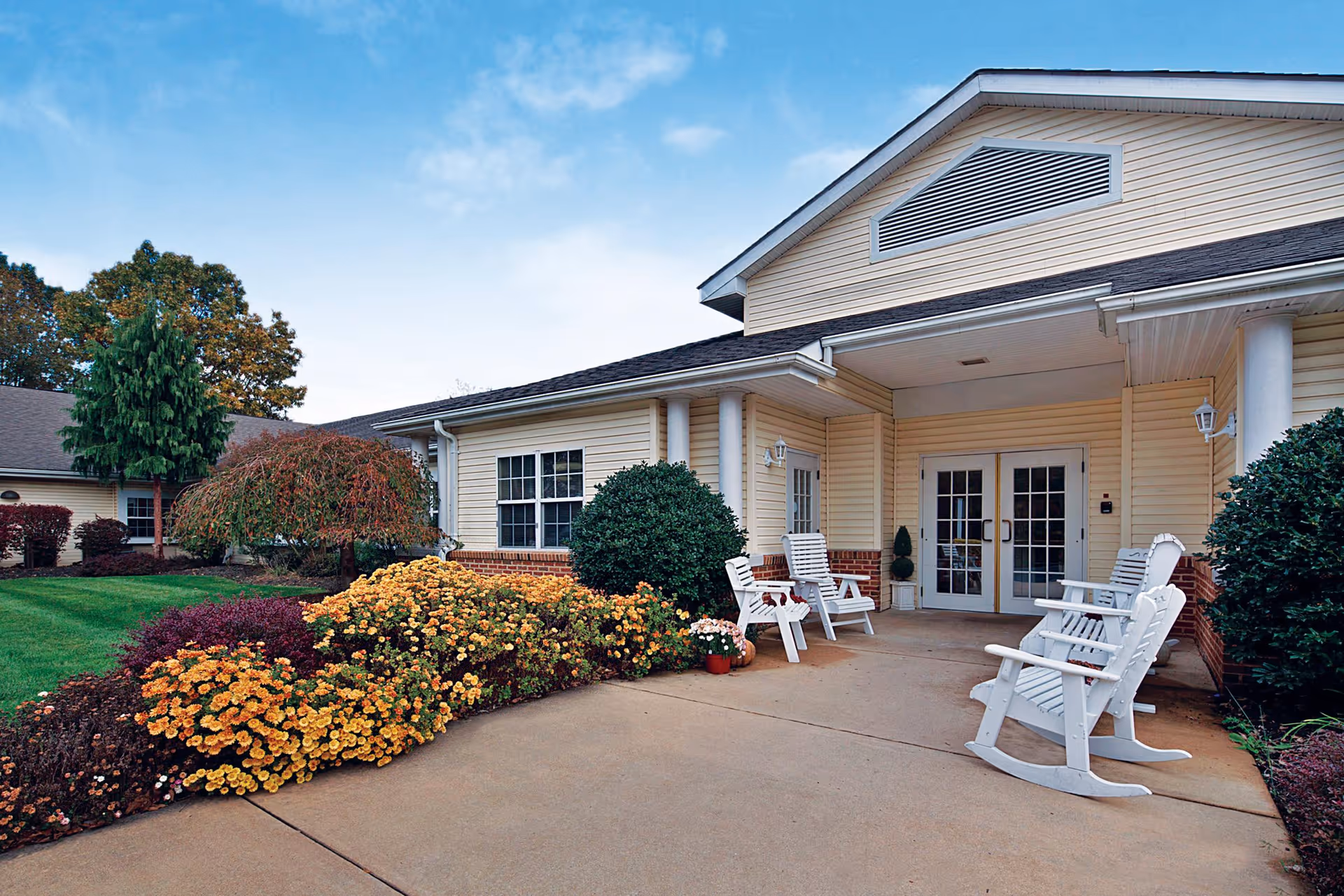 Front entrance of a light-colored building with a covered porch, white rocking chairs, and colorful flowerbeds.
