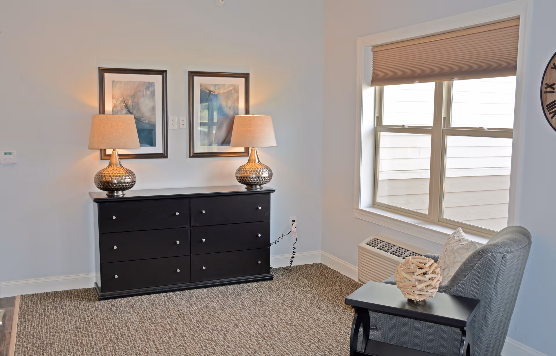A cozy interior corner of a room featuring a black dresser with six drawers, topped with two matching table lamps and two framed abstract paintings on the wall above. To the right, there is a window with beige blinds partially drawn, an air conditioning unit below it, and a gray upholstered chair next to a small black side table holding a decorative wooden sphere.