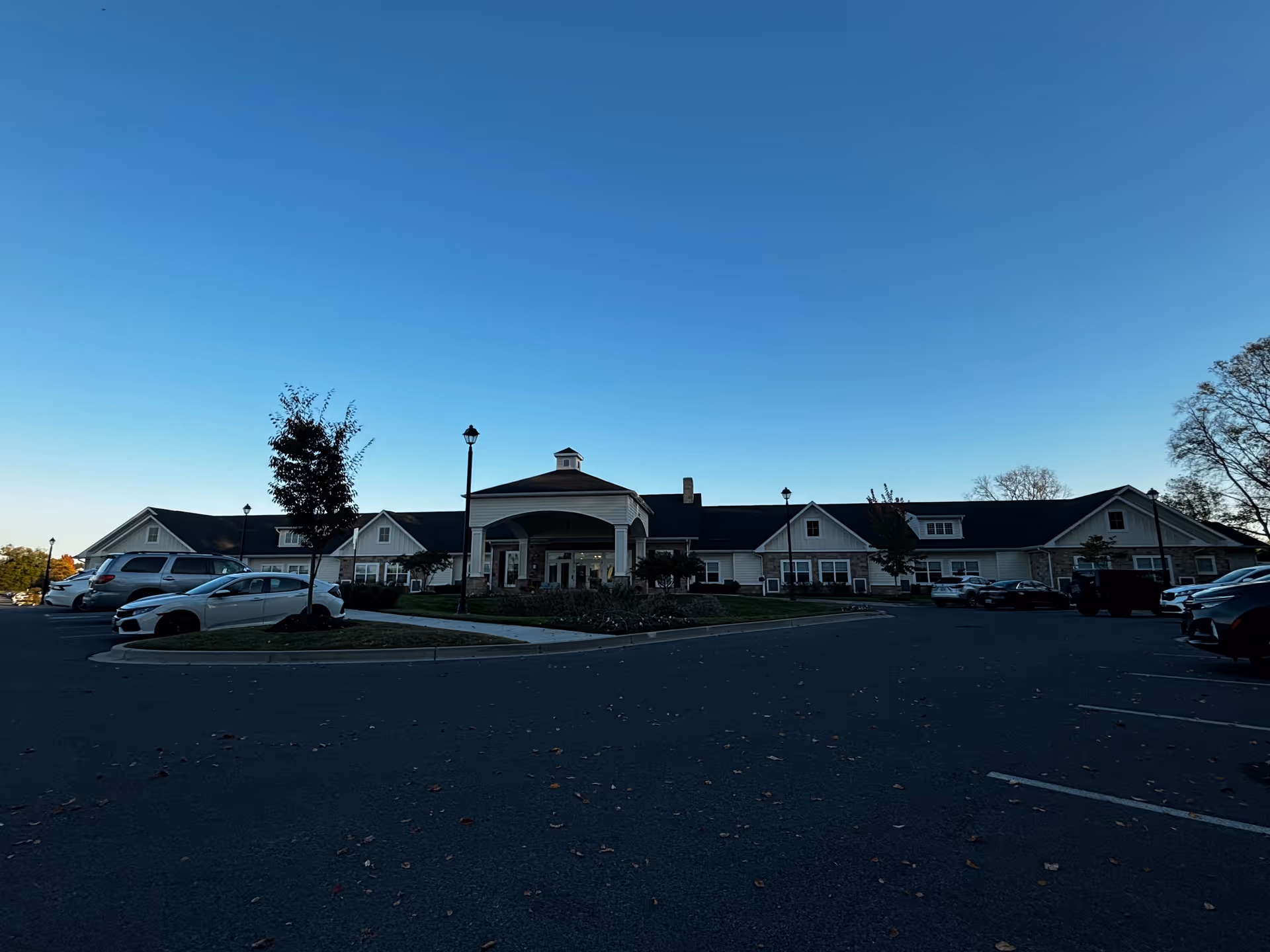 Wide exterior view of Silverado Frederick Memory Care Community building with a large parking lot in front, several parked cars, a clear blue sky, and some trees around the property.