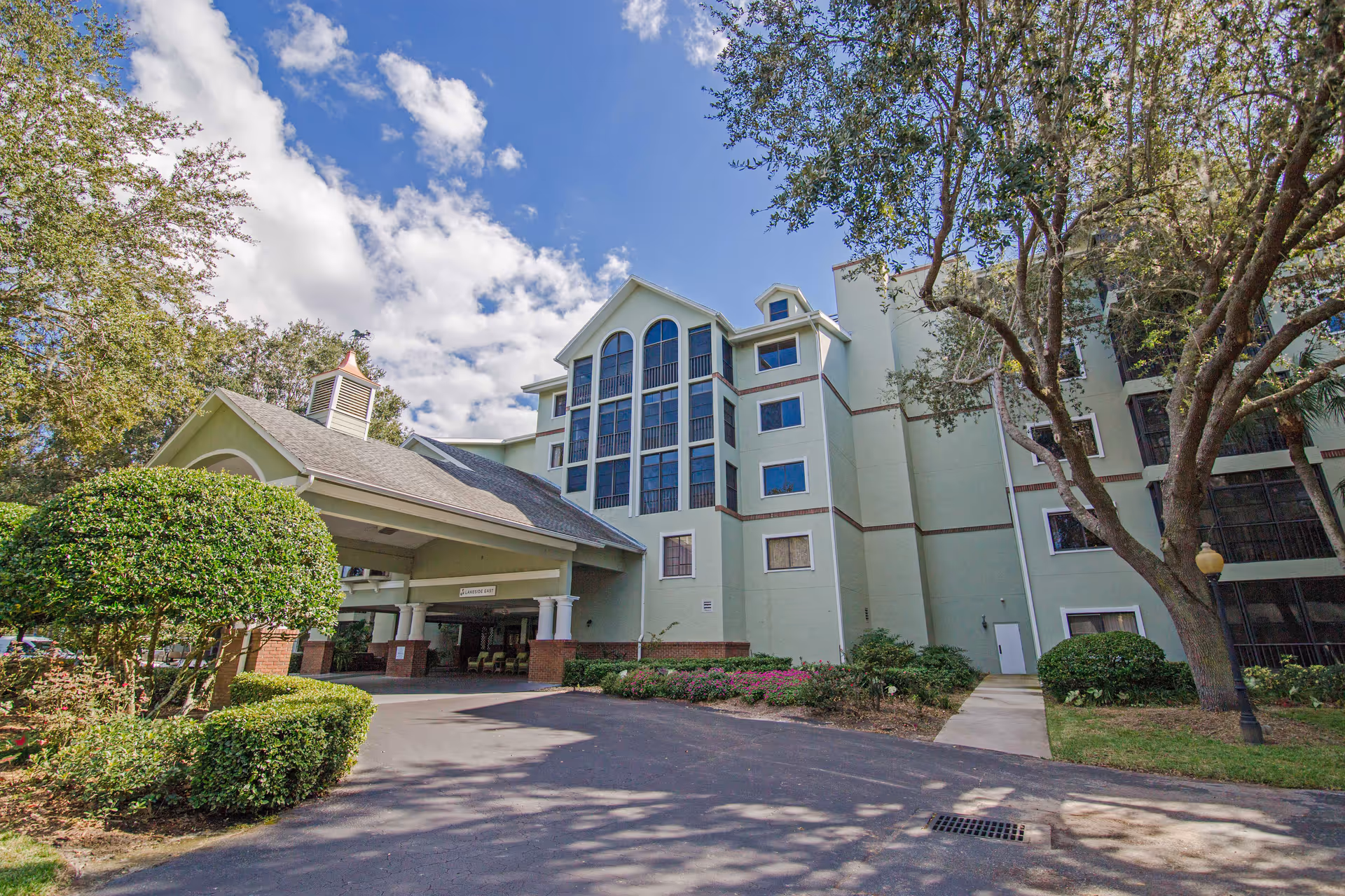 Exterior view of a multi-story senior living facility building with a covered entrance, surrounded by trees and landscaped bushes under a partly cloudy sky.