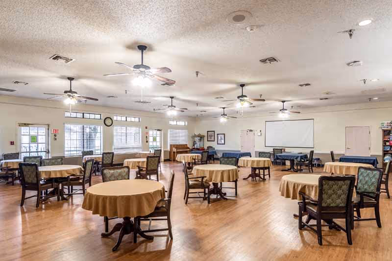 A spacious room with multiple round tables covered with beige tablecloths, each surrounded by wooden chairs with patterned cushions. The room has wooden flooring, several ceiling fans with lights, large windows letting in natural light, and a piano against one wall. There is also a projection screen on the far wall and some framed pictures and shelves.