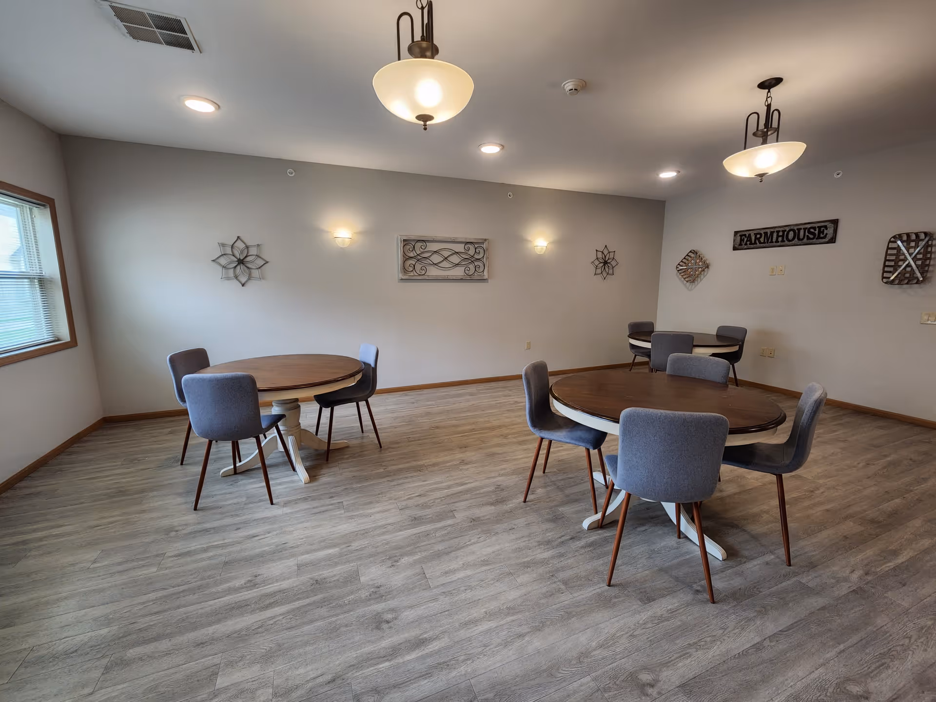 A dining area with three round wooden tables, each surrounded by four gray upholstered chairs. The room has light gray walls decorated with metal wall art and a sign that reads 'FARMHOUSE'. The floor is covered with light wood laminate, and there are two ceiling light fixtures providing warm lighting. A window on the left side lets in natural light.