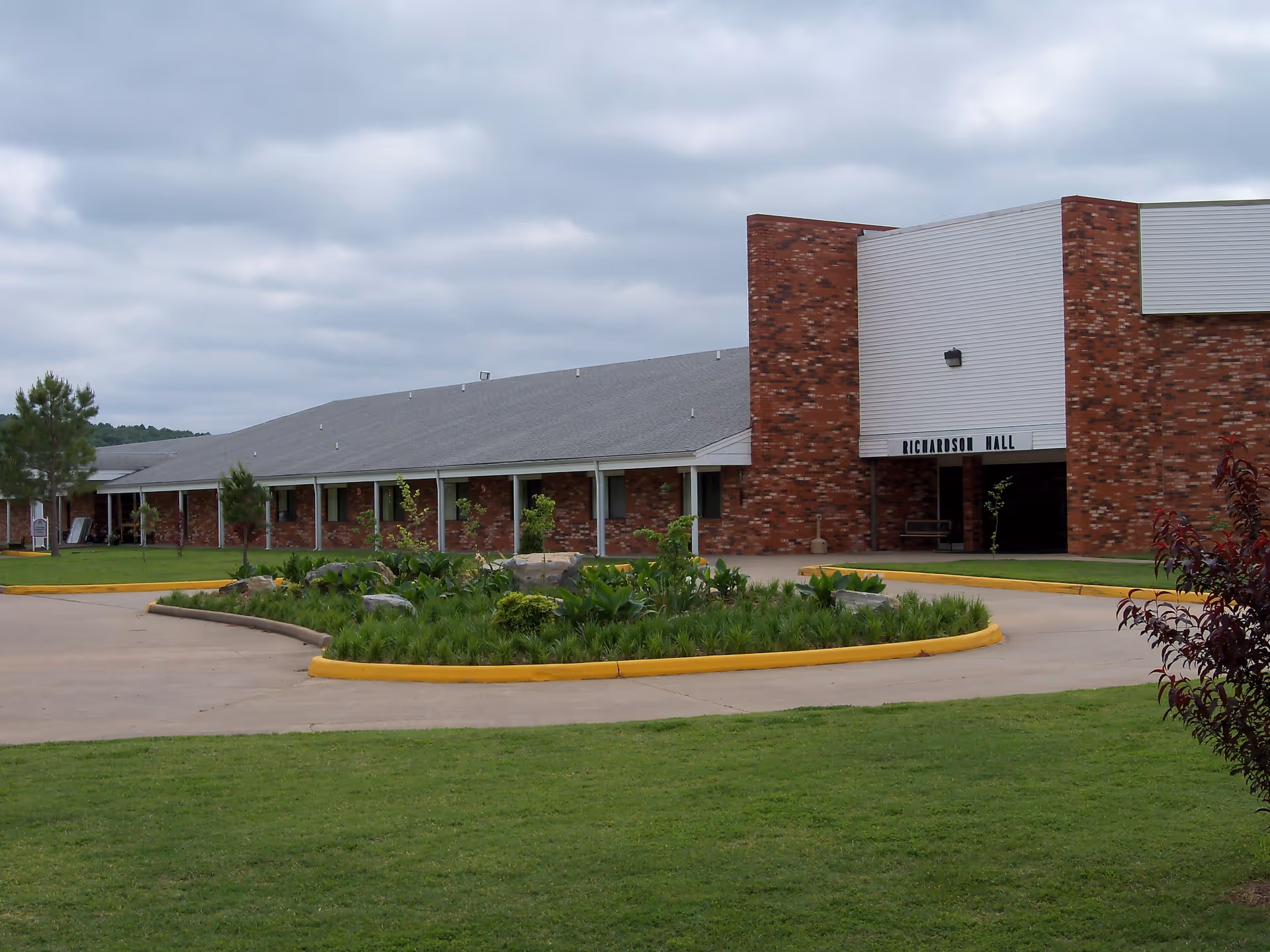 Exterior view of a single-story brick building named Richardson Hall with a gray roof, surrounded by a well-maintained lawn and a circular driveway with a landscaped island in the center. The sky is cloudy.