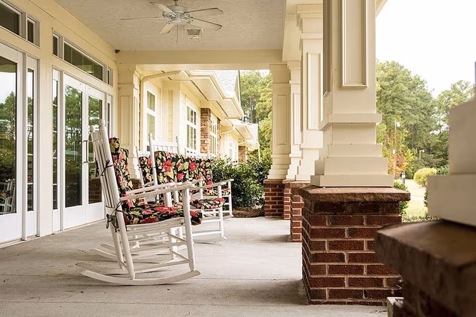A covered outdoor porch area with white rocking chairs that have floral cushions. The porch has large white columns with brick bases and overlooks a green garden area with trees.