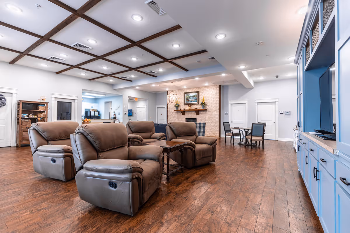 Spacious living room area with four brown recliner chairs arranged around a wooden coffee table. The room features wood flooring, a ceiling with exposed wooden beams and recessed lighting, a brick fireplace with a painting and floral decorations above it, and a blue built-in entertainment center with a TV. In the background, there is a small dining table with four chairs and a view into a kitchen area.