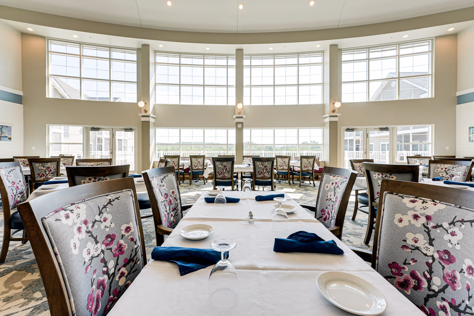 Bright dining room with large windows allowing natural light to fill the space. Tables are set with white tablecloths, plates, upside-down glasses, and navy blue napkins. Chairs have floral patterned upholstery. The room has a high ceiling and a view of the outdoors through the windows.