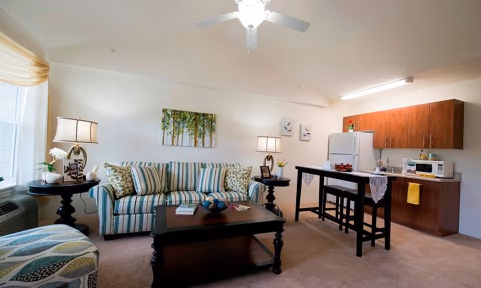A cozy living area with a striped sofa adorned with patterned cushions, two side tables each with a lamp, a coffee table with decorative items, and a small kitchen area in the background featuring wooden cabinets, a refrigerator, microwave, and a high table with two stools. The room is well-lit with natural light from a window and a ceiling fan with a light fixture.