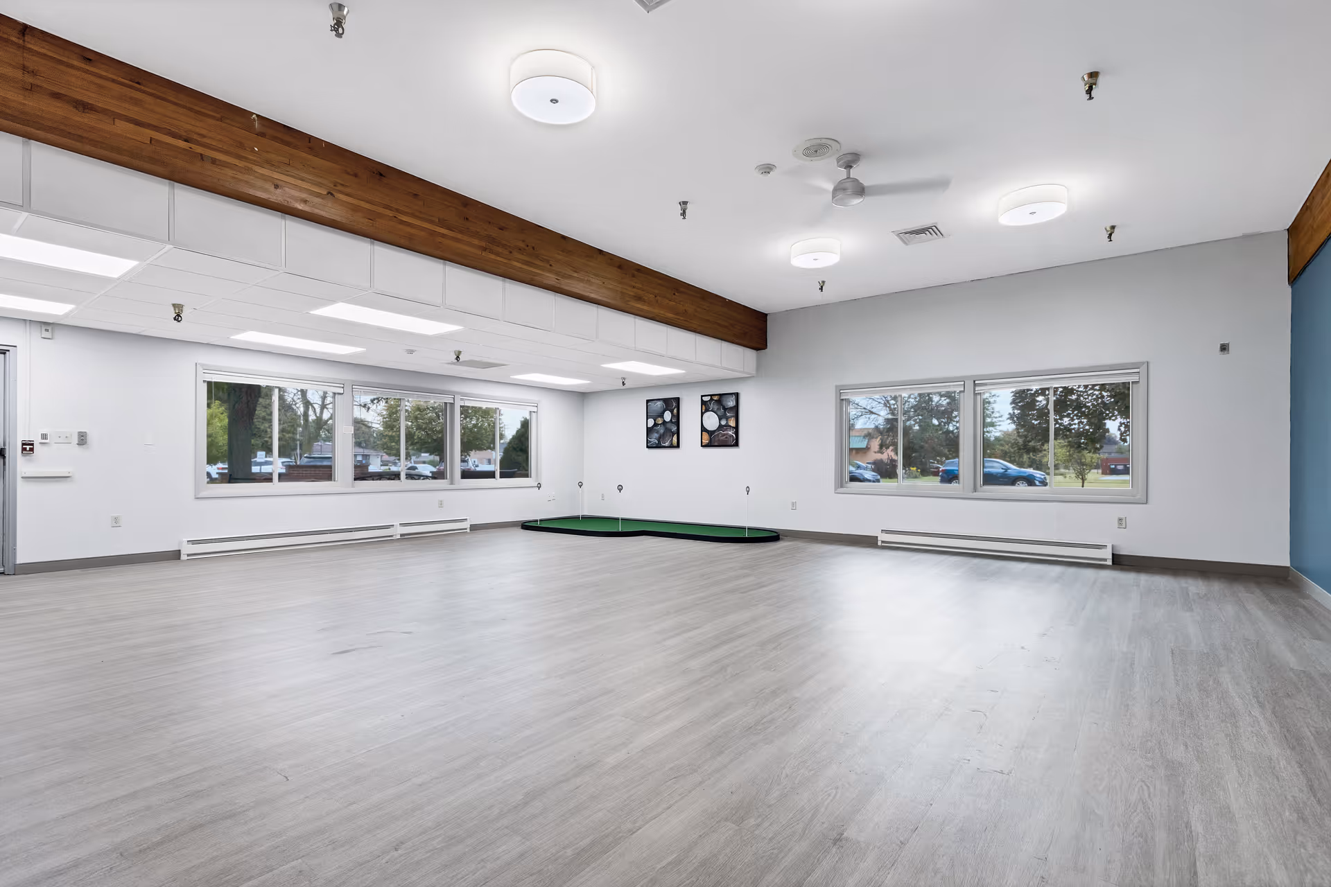 Spacious empty multipurpose activity room with large windows, a wood beam, ceiling lights, and light wood flooring.