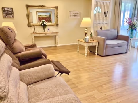 A cozy living room with beige reclining chairs on the left and a gray armchair on the right. The room has wooden flooring and light-colored walls. There is a small wooden side table with a lamp and tissue box, a console table with a vase of flowers, and a large ornate mirror above it. Wall decor includes a sign that says 'Together'. A sliding glass door with vertical blinds leads to an outdoor area with visible greenery.