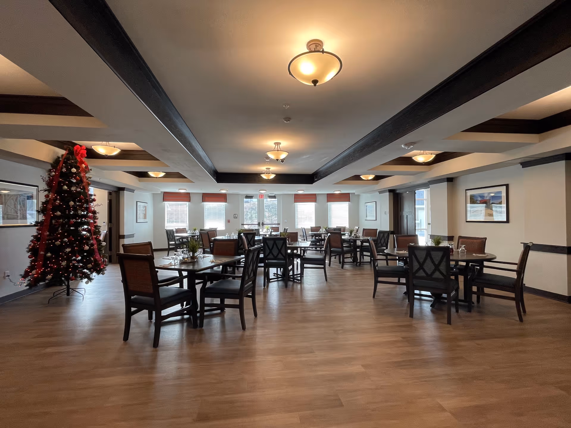 A spacious dining room with multiple tables and chairs arranged neatly. The room features wooden flooring, white walls with dark wood trim, and ceiling lights. A decorated Christmas tree with red ribbons and ornaments stands on the left side near the wall. Several windows with red valances allow natural light to enter the room.