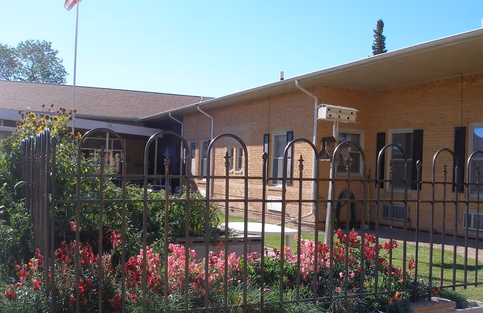 Outdoor garden area at Mondell Heights Retirement Community featuring a black metal fence, colorful flowers, green shrubs, a birdhouse on a pole, and a brick building with windows and a covered walkway in the background under a clear blue sky.