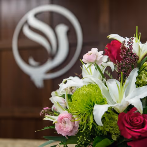 Close-up of a colorful floral arrangement with white lilies, pink and red roses, and green chrysanthemums in front of a wooden wall featuring a blurred circular logo.