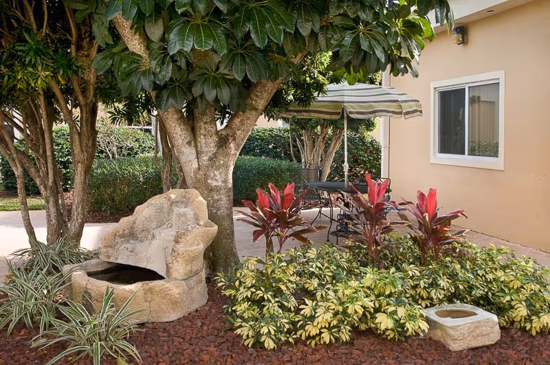 Outdoor courtyard area with a stone fountain, trees, a patio table with umbrella, and landscaped plants beside a beige building.