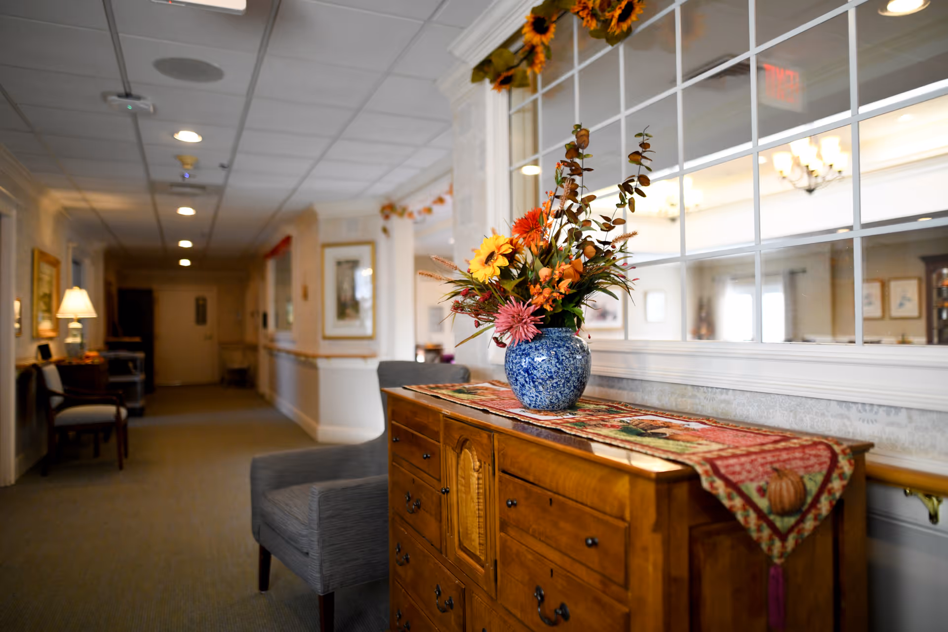 A well-lit hallway in an assisted living facility with a wooden sideboard topped with a colorful floral arrangement in a blue vase. The hallway has framed artwork on the walls, a gray upholstered chair, and a large window with multiple panes reflecting a chandelier and other interior furnishings.