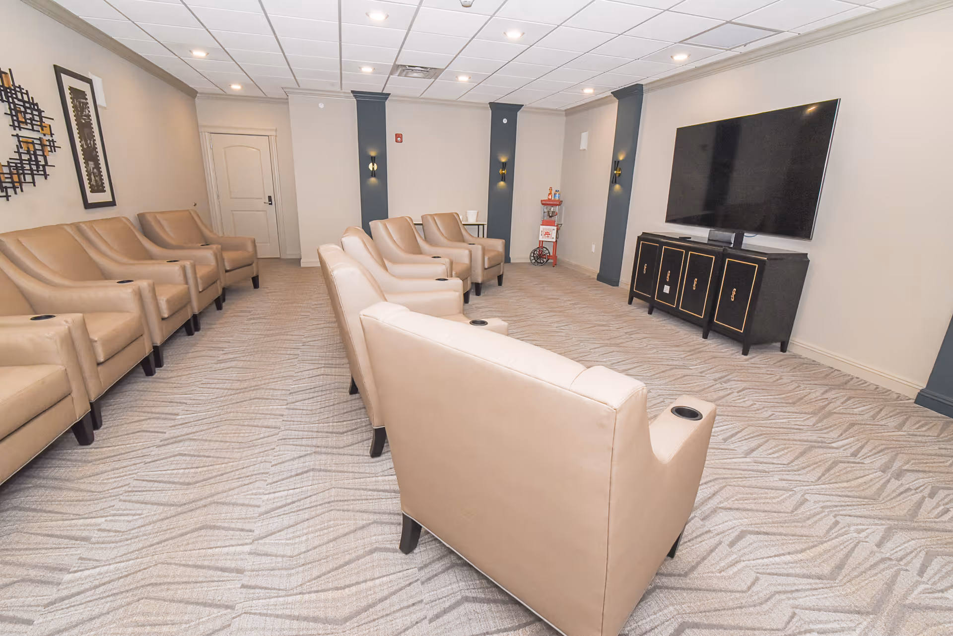 A room with two rows of beige leather armchairs facing a large flat-screen TV mounted on the wall. The room has light-colored walls, patterned carpet flooring, and decorative wall sconces. There is a popcorn machine in the corner and framed artwork on one wall.