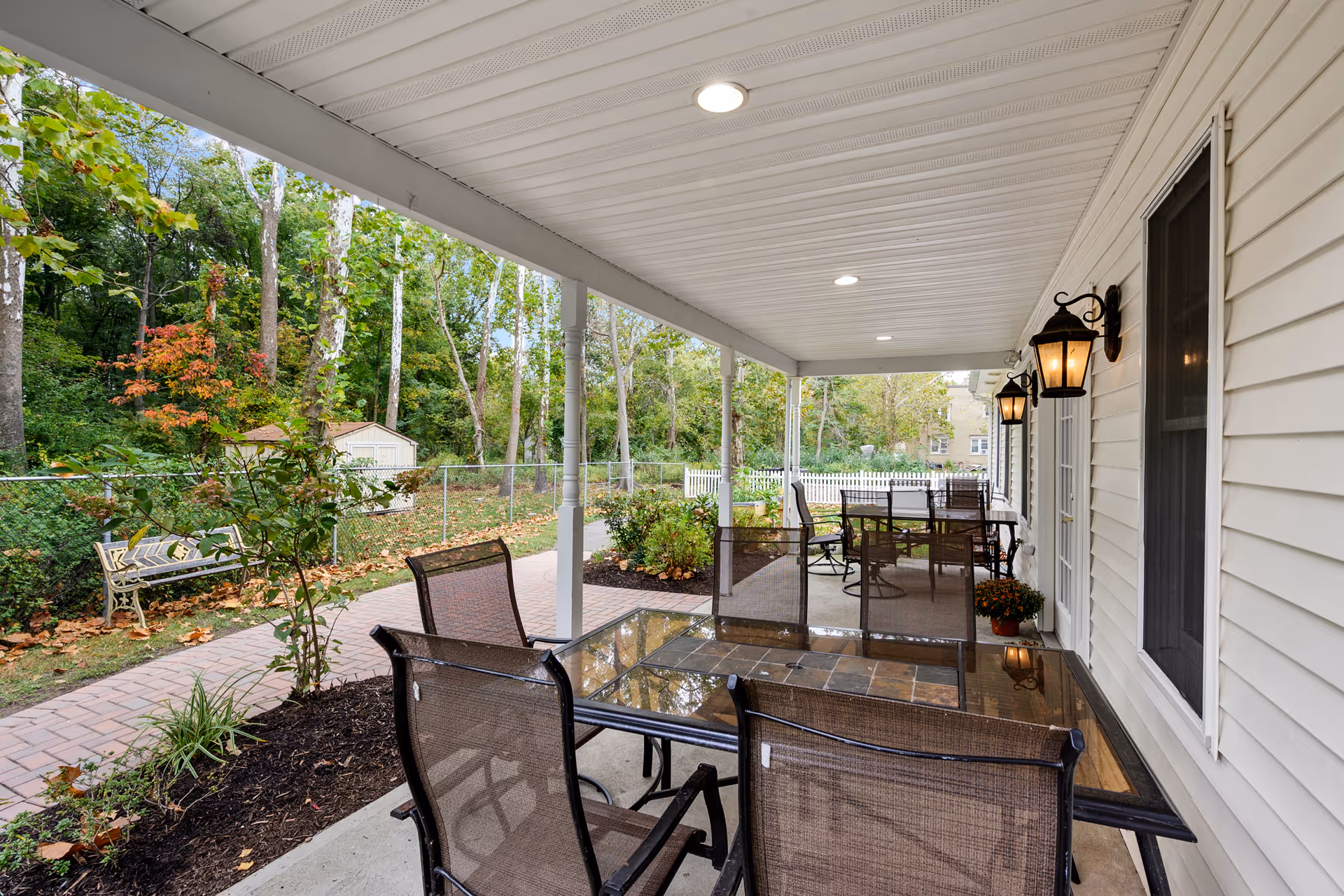 Covered patio with glass-top tables and chairs overlooking a landscaped yard, walkway, and trees.