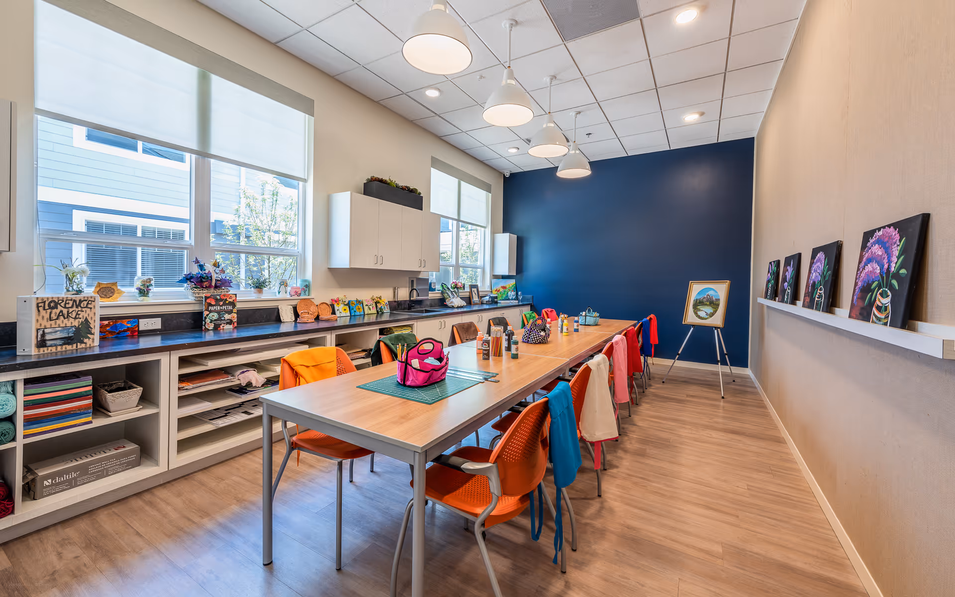 A bright and spacious craft room with a long table surrounded by orange chairs, some with colorful jackets draped over them. The table has various craft supplies and a pink bag on it. The room features large windows with white blinds, white cabinets, and shelves filled with art materials. The far wall is painted dark blue and displays framed artwork on a ledge and an easel with a painting.