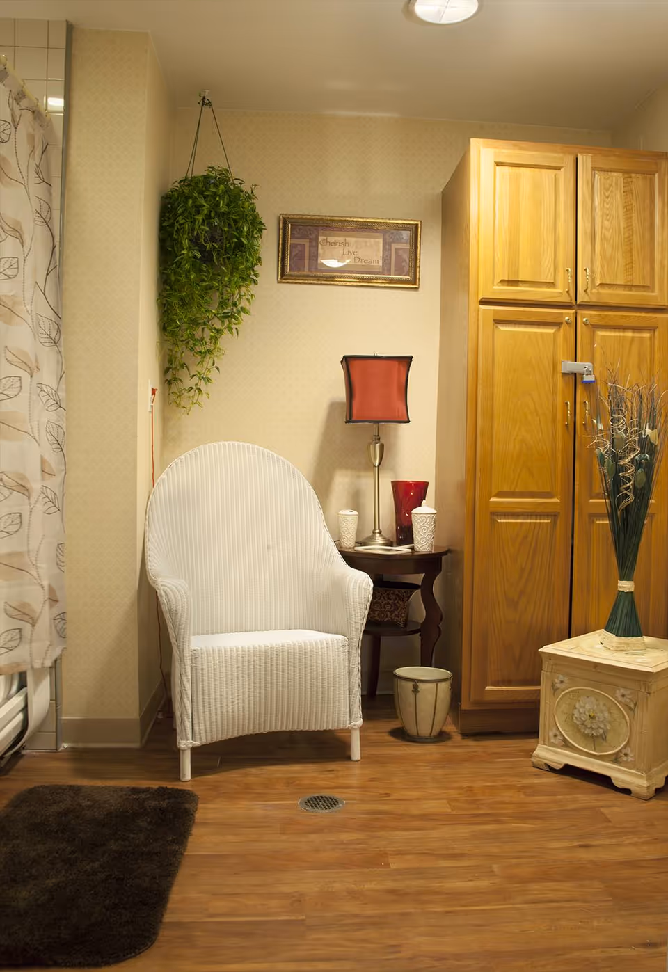 A cozy corner of a room featuring a white wicker chair, a small wooden side table with a red lampshade and decorative items, a tall wooden cabinet, a hanging green plant, a framed inspirational picture on the wall, and a decorative vase on a small wooden stand. The floor is wooden with a dark brown rug partially visible.