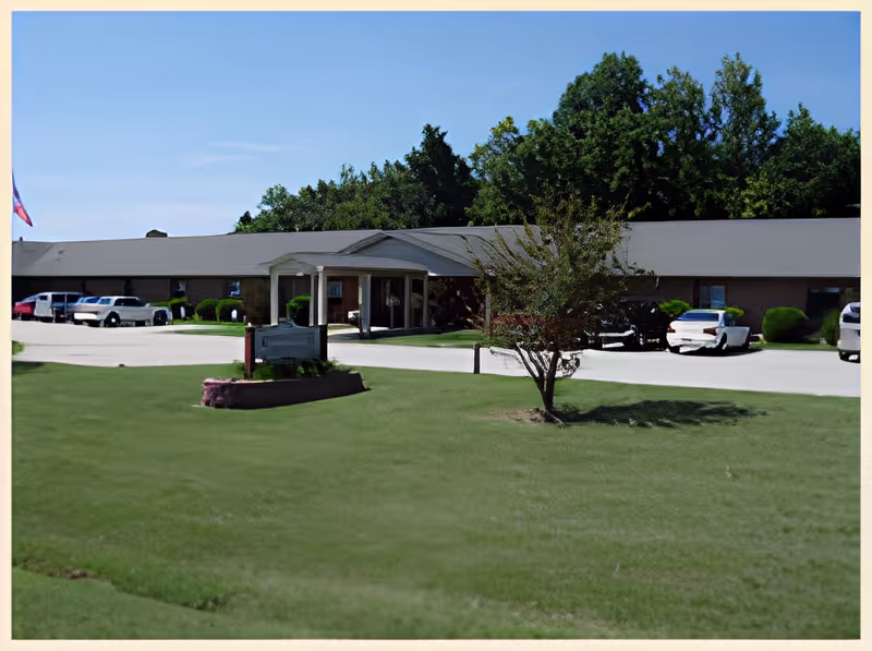 Exterior view of Brookstone Retirement Center showing a single-story brick building with a covered entrance, several parked cars, a green lawn with a small tree, and a flagpole with a flag on the left side.