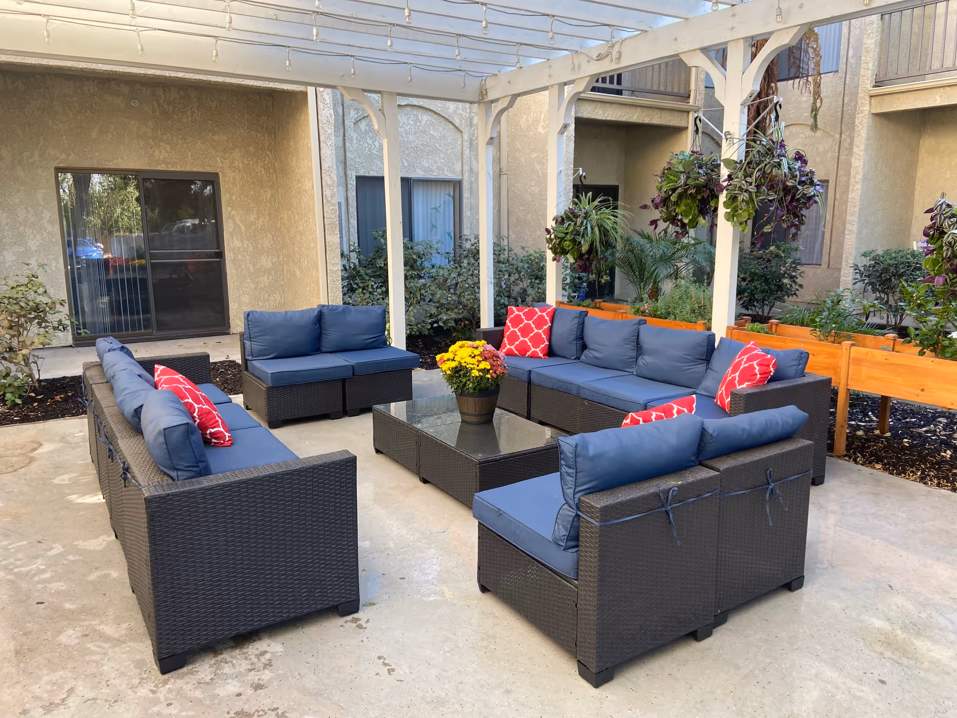 Outdoor patio area with dark wicker furniture arranged in a U-shape under a white pergola. The seating has blue cushions and red patterned throw pillows. A glass-top coffee table with a potted yellow flower is in the center. There are hanging plants and raised garden beds with greenery around the patio, adjacent to a beige building with windows and sliding glass doors.