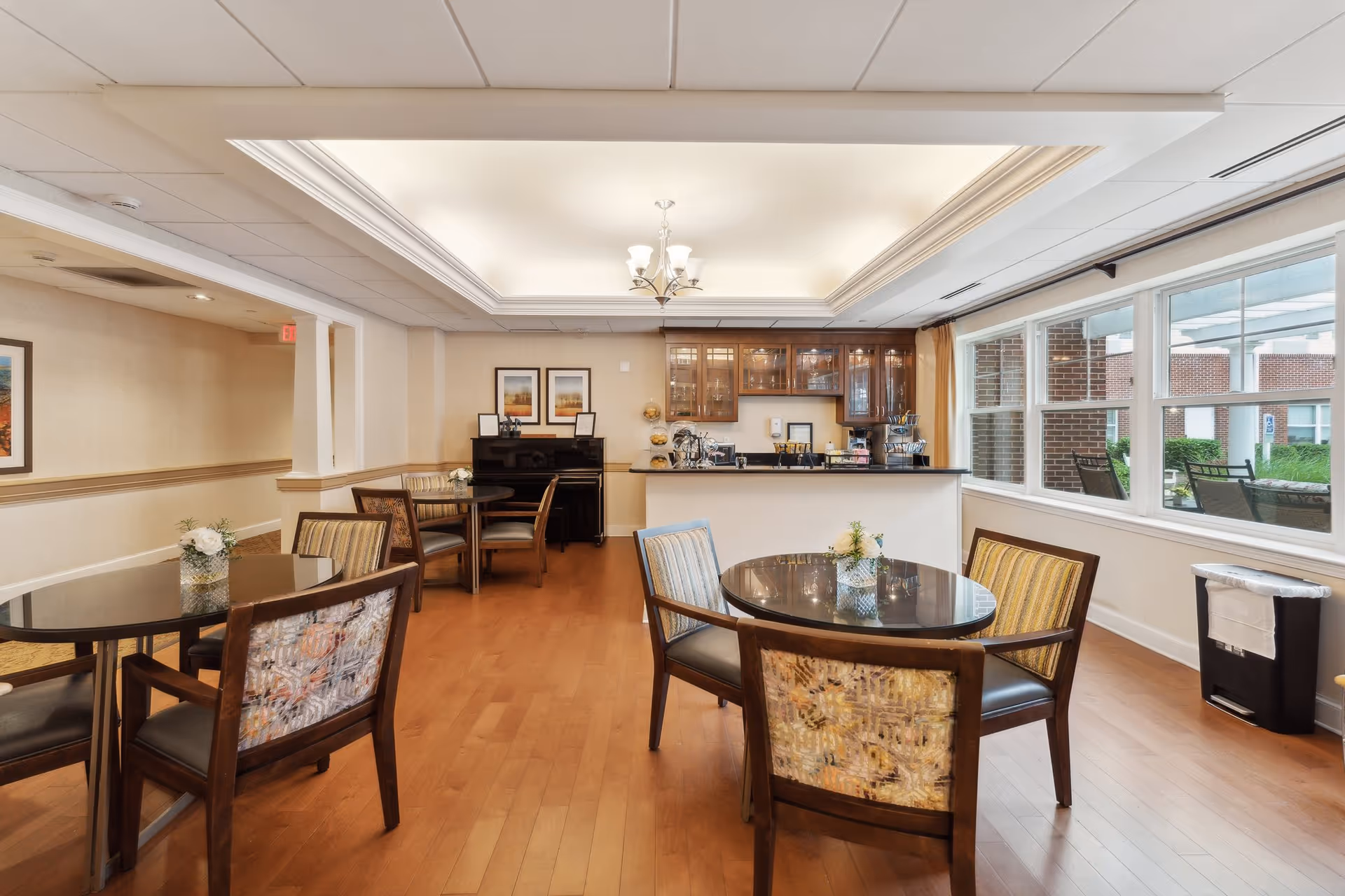 A bright and spacious dining area with several round tables and cushioned chairs. The room features wooden flooring, a ceiling light fixture, large windows letting in natural light, and a counter with glass cabinets above it. There is a piano against the far wall and framed artwork hanging above it. Outside the windows, a patio area with chairs is visible.